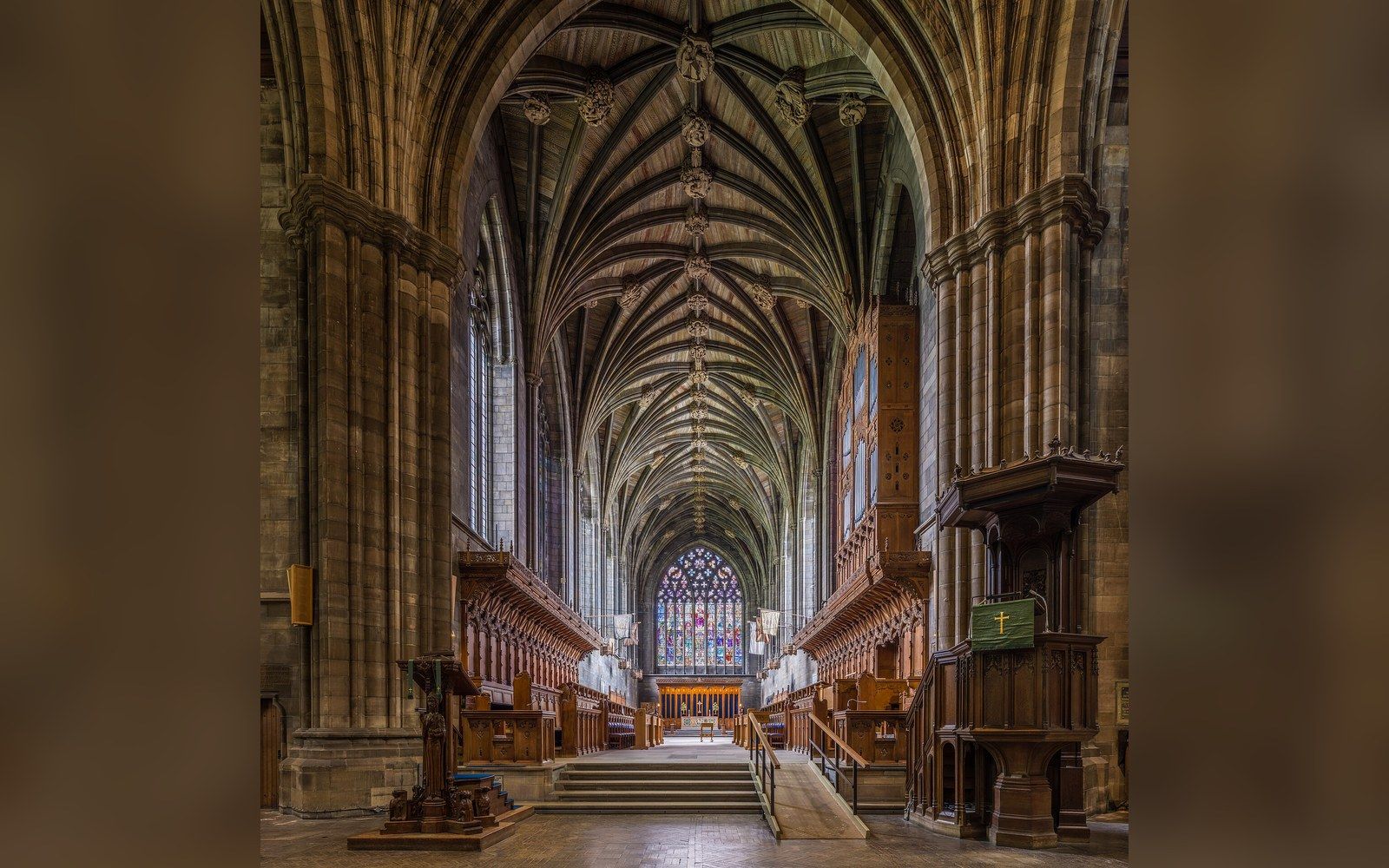 Vaulted stone ceiling and choir stalls lead toward a stained glass window in an abbey