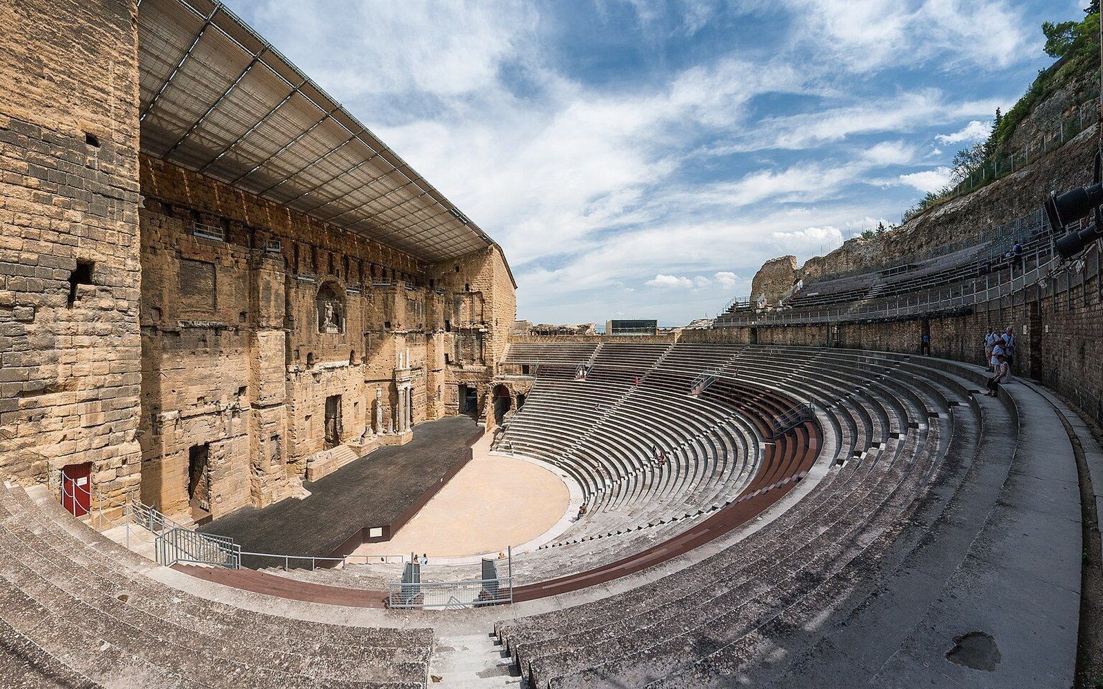 Curved stone seating tiers face the ancient theatre's towering stage wall under a bright sky